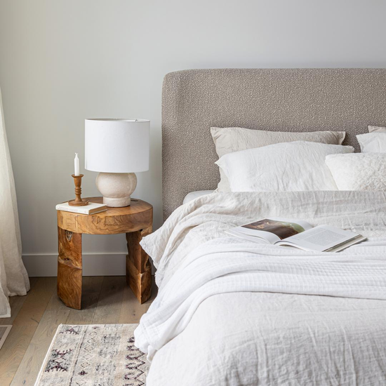 Natura Bundar Accent Table in Natural teak beside an upholstered bed
